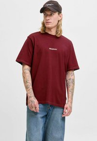 Bordo lielais T-krekls ar uzrakstu "ORIGINALS" uz krūtīm. Kokvilnas audums, īsas piedurknes, apaļa kakla izgriezums, savienots ar gaiši ziliem džinsiem.