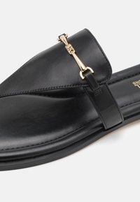 Sandalo scivolato in pelle nera con una fascia fissata da accessori in tono oro; texture liscia e design minimalista. Presenta una suola piatta.