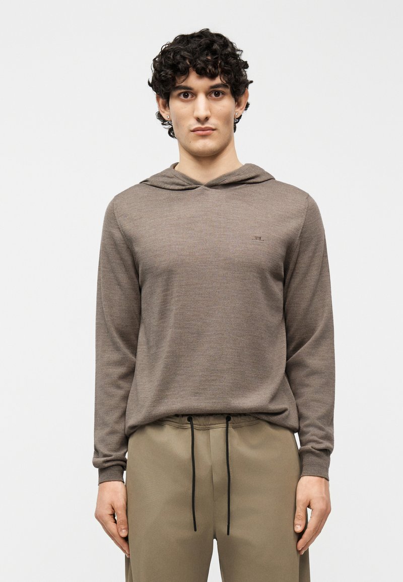 Νέος με σγουρά μαλλιά, φορώντας καφέ φούτερ με κουκούλα και μπεζ παντελόνι με κορδόνι, στέκεται μπροστά σε απλό φόντο.