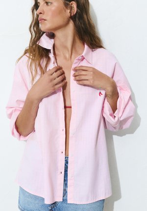 Frau mit langen Haaren richtet ein offenes hellrosa Hemd mit Chili-Stickerei, trägt blaue Jeans und rote Unterwäsche-Träger sind sichtbar.