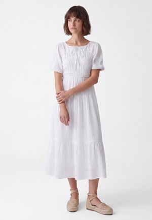 Woman with short brown hair stands wearing a white midi dress with short puff sleeves and beige espadrille wedges.