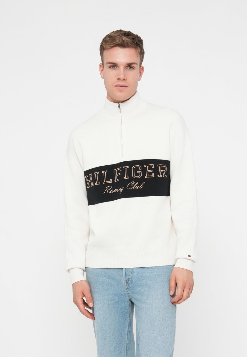 Balta zip-up džemperis ar melnu horizontālu joslu, uz kuras rakstīts "HILFIGER Racing Club" zeltā. Saskaņots ar gaiši zilām džinsām. Mīksta tekstūra.