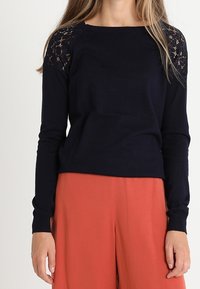 Žena oblečená v černém dlouhém rukávu s krajkovými rameny a širokých kalhotách v barvě rzi, stojící proti jednolitému pozadí.