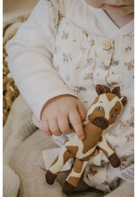 Soft plush toy shaped like a giraffe, featuring patterned brown and cream fabric, button accents, and a smiling face. Child's hand holds it.