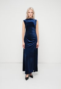 Γυναίκα με μακριά ξανθά μαλλιά που φοράει ένα αμάνικο βελούδινο φόρεμα μέχρι το πάτωμα σε ναυτικό μπλε χρώμα και μαύρες μυτερές γόβες, στολισμένη μπροστά σε λευκό φόντο.