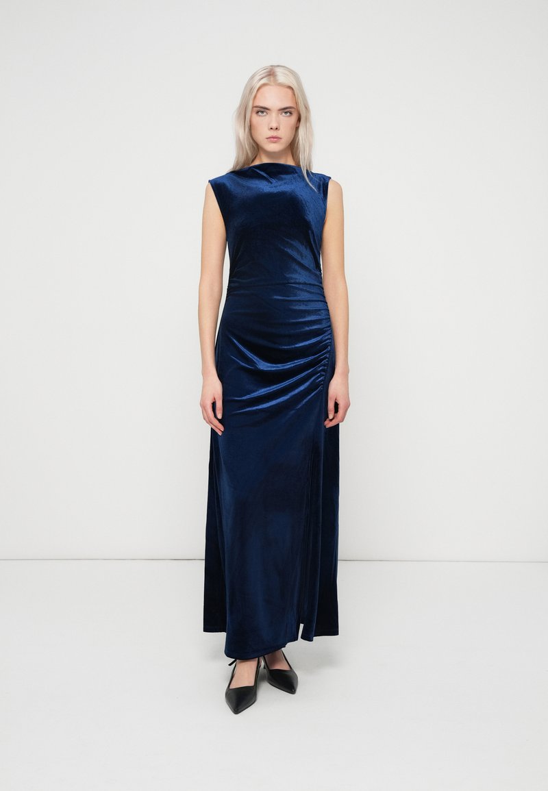 Γυναίκα με μακριά ξανθά μαλλιά που φοράει ένα αμάνικο βελούδινο φόρεμα μέχρι το πάτωμα σε ναυτικό μπλε χρώμα και μαύρες μυτερές γόβες, στολισμένη μπροστά σε λευκό φόντο.