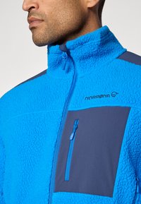 Giacca in pile di un blu vivace con tessuto testurizzato, colletto alto, tasca sul petto con zip, pannellatura blu navy e zip a contrasto.