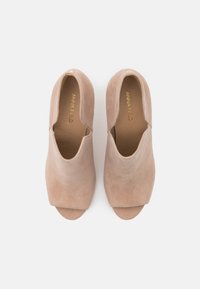 Mule beige in suede con punta aperta, dalla texture liscia, bordi arrotondati e cuciture dettagliate. L'interno presenta il nome del marchio in oro.