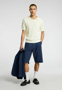 Polo crema a maniche corte con texture a coste, pantaloni corti blu navy, mocassini neri e calzini bianchi; tiene una giacca blu navy. Outfit semplice e strutturato.