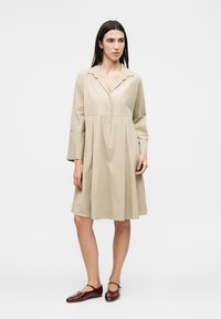 Γυναίκα με ίσια σκούρα μαλλιά που φοράει χαλαρό μπεζ μακρυμάνικο φόρεμα πουμοκαμπά και σκούρα κόκκινα ψηλοτάκουνα παπούτσια, στέκεται μπροστά σε ένα απλό φωτεινό φόντο.