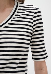 Eine Frau trägt ein kurzärmliges, schwarz-weiß geringeltes Rippoberteil mit V-Ausschnitt, zu sehen von Hals bis zur Mitte des Oberkörpers.