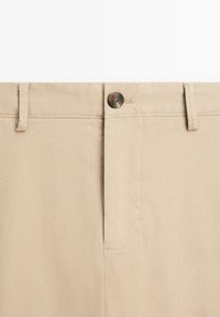 Beige cotton trousers with a zipper fly and a single brown button. Features a flat front, belt loops, and side pockets. Smooth texture.