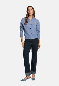 Donna che indossa un cardigan azzurro chiaro con bottoni e un emblema, jeans scuri con risvolti e scarpe slingback basse coordinate di colore azzurro chiaro, in piedi.
