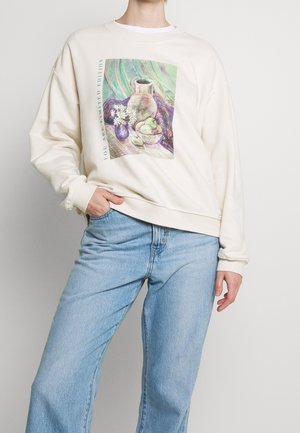 Person trägt einen cremefarbenen Sweatshirt mit pastellfarbenem Stillleben-Druck von Vase, Blumen und Obst, kombiniert mit hellblauen Jeans, Hand in der Tasche.