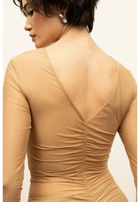 Nude bodysuit z długim rękawem i V-kształtnym wycięciem z tyłu. Gładka faktura, marszczenia po bokach. Srebrna kolczyk widoczny w profilu.