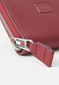 Raudona tekstūruota odinė piniginė su sidabriniu užtrauktuku. Yra logotipo etiketė su užrašu "TOMMY JEANS." Detaliu vaizdas, akcentuojantis užtrauktuko traukiklio ir medžiagos ypatybes.