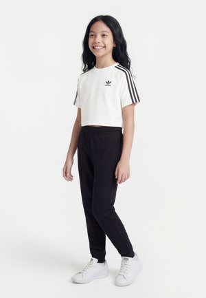 Fille souriante debout portant un crop top Adidas blanc avec des bandes noires, un pantalon de jogging noir et des baskets blanches sur un fond blanc uni.