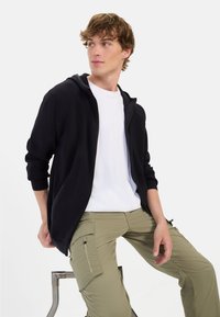 Schwarze Zip-Up-Byjacke mit gerippter Textur über einem weißen T-Shirt, kombiniert mit hellgrünen Cargo-Hosen mit Taschen und Reißverschlüssen.
