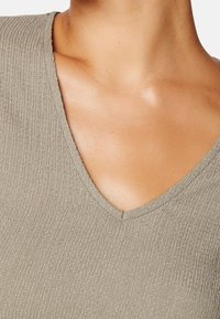 Primo piano del collo di una persona che indossa un top beige a V con texture, che mostra una pelle liscia e clavicole.