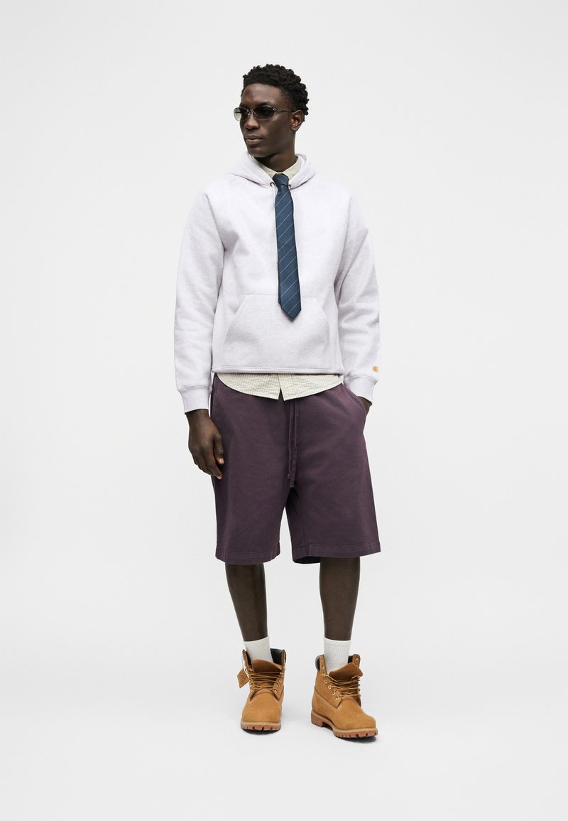 Man met een zonnebril, witte hoodie over een overhemd met een blauwe stropdas, paarse korte broek, witte sokken en beige veterlaarzen, staand met handen in de zakken.