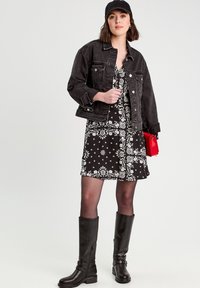 Veste en denim noire sur une robe fleurie noir et blanc, assortie à des bottes noires montantes et un sac à main rouge. Le look présente de forts contrastes.