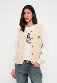 Μπεζ γιλέκο από φλις με στρογγυλή λαιμόκοψη, πέντε καφέ κουμπιά και λεπτομέρεια με λεοπάρ καρδιά. Φορεμένο πάνω από ένα άσπρο φούτερ και μπλε τζιν.