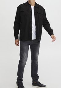 Zwart shirtjack met twee voorzakken, gecombineerd met een wit T-shirt en donkergrijze spijkerbroek. Zwarte sneakers maken de look compleet.