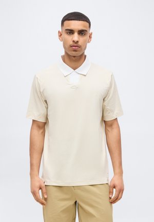 Noor mees lühikeste juustega, kandes beežikat lühikeste varrukatega särki valge kraelise särgi kohal, seisab ühevärvilise heleda tausta ees.