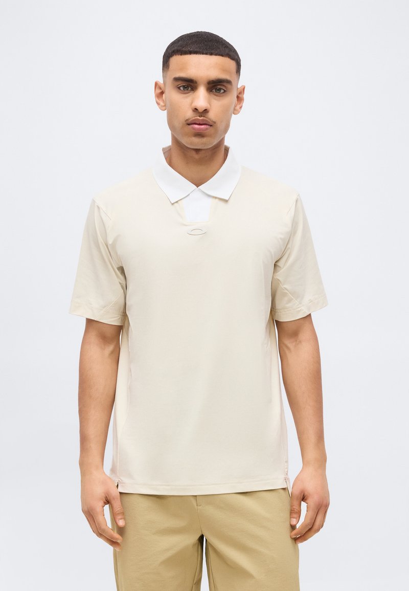 Νέος άνδρας με κοντά μαλλιά, φοράει μπέζ πουκάμισο με κοντά μανίκια πάνω από λευκό πουκάμισο με γιακά, στέκεται μπροστά σε απλό ανοιχτό φόντο.