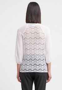 Woman with straight dark hair wears white blouse with lace back and sheer sleeves, paired with black pants, standing against plain background.