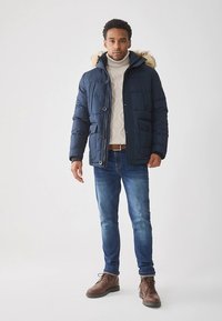 Mörkblå vadderad jacka med pälskantad huva, quilteffekt och flera fickor, använd över en krämfärgad kabelstickad tröja och blå jeans.