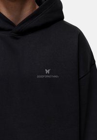 Sudadera negra con un pequeño logo de mariposa gris sobre el texto "GOODFORNOTHING" en el pecho, llevada por una persona.