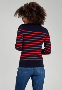 Maglione blu navy con strisce orizzontali rosse, design aderente, maniche lunghe e colletto a costine. Indossato con jeans in denim di lavaggio medio.