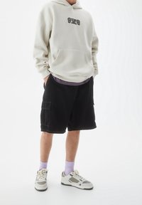 Sudadera color beige claro con gráfico en el frontal, combinada con pantalones cortos cargo negros con bolsillos laterales. Zapatillas gris y blancas con calcetines morados.
