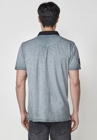 Siva polo majica s črnimi ovratnikom, kratkimi rokavi in zbledelim ombre učinkom. Ima logotip na levem ramenu. Gladka tekstura.