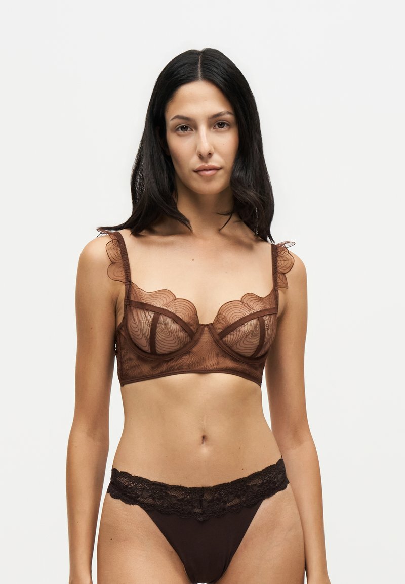 Femme aux longs cheveux foncés portant un soutien-gorge en dentelle marron transparent et une culotte assortie bordée de dentelle sur un fond clair uni.