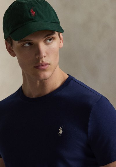 Μπλε μπλούζα από βαμβάκι με στενή εφαρμογή και μικρό ασημένιο λογότυπο polo στο στήθος· συνδυάζεται με σκούρο πράσινο καπέλο μπέιζμπολ με κόκκινο λογότυπο.