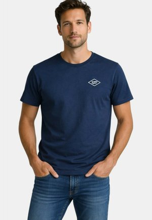 Homme portant un t-shirt bleu marine Lee Cooper et un jean bleu, debout les mains dans les poches contre un fond blanc.