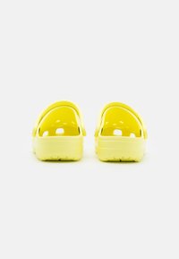 Um par de tamancos amarelos brilhantes, estilo slip-on, com buracos de ventilação, visto por trás sobre um fundo branco.