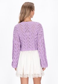 Pull en tricot violet avec un motif en chevrons, manches longues et design cropped. Porté par-dessus une robe blanche ornée de dentelle.