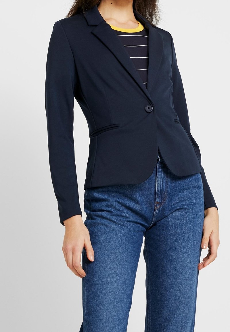Námořnický blazer s přiléhavým střihem, zapínáním na jeden knoflík a dvěma předními kapsami. Nesený na námořnickém pruhovaném tričku a modrých džínách.