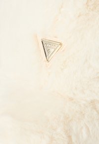 Logo triangulaire doré de la marque Guess sur un tissu en peluche doux de couleur crème, mettant en valeur la texture et les détails de la marque.