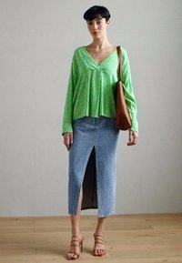 Grønn langermet topp med V-hals, iført en lys blå denimskjørt med splitt foran. Brun veske og sandaler med stropper fullfører antrekket.