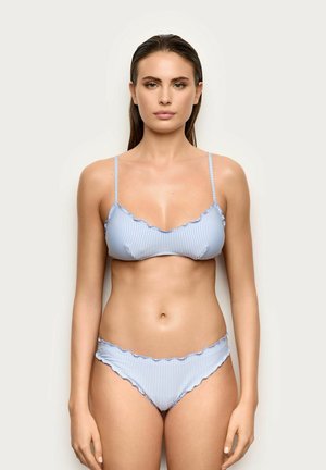 Kobieta w jasno-niebieskim i białym bikini w paski z falistymi krawędziami, stojąca na jednolitym jasnym tle, zwrócona przodem.