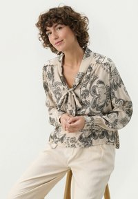 Krämfärgad blus med svart paisley-mönster, knytkrage, långa ärmar och knappdetaljer. Matchas med ljusbeiga byxor.