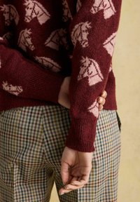 Person in einem bordeauxfarbenen Pullover mit rosa Pferdeköpfen und karierten Hosen, der mit einer Hand den gegenüberliegenden Arm hinter dem Rücken hält.