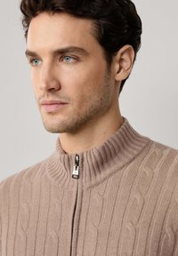 Beżowy sweter zapinany na zamek z prążkowanym kołnierzem, z charakterystycznym wzorem w warkocze. Zamek błyskawiczny w kolorze srebrnym wyróżnia się na tle materiału.