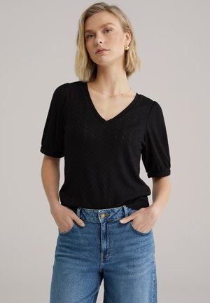 Haut noir à col en V avec un motif chevron texturé et des manches bouffantes, associé à un jean bleu taille haute et un collier en or.