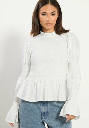 Frau mit glattem dunklem Haar, die eine weiße geraffte Bluse mit Puffärmeln und Glockenmanschetten trägt, kombiniert mit blauen Denim-Jeans.