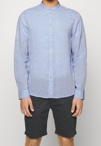 Camicia a maniche lunghe blu chiaro con colletto alla francese, design con bottoni davanti e tessuto morbido e texturizzato. Abbinata a pantaloni corti scuri.